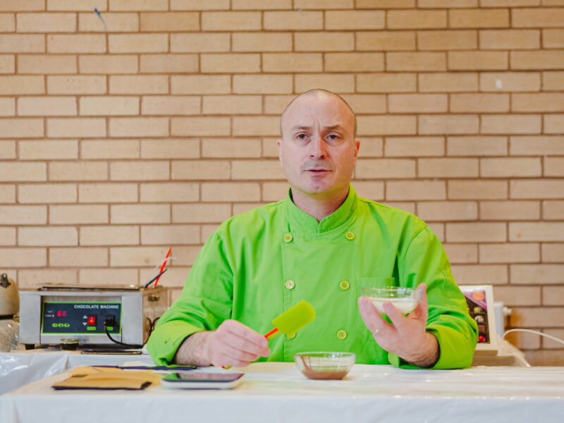 A man in a green chefs apron is sitting at a table combining ingredients in small glass bowls